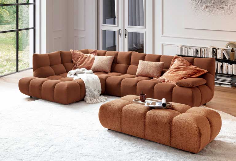 Große modulare Eckcouch von 3C Candy in Rostbraun mit Würfelsteppung, platziert auf einem weißen Hochflorteppich in einem hellen, modernen Wohnzimmer.